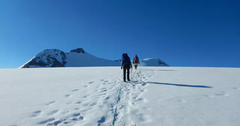 Twee bergbeklimmers met rugzakken lopen achter elkaar over een besneeuwde, zonovergoten berghelling onder een strakblauwe hemel, terwijl ze voetafdrukken achterlaten. Op de achtergrond zijn met sneeuw bedekte bergtoppen te zien.